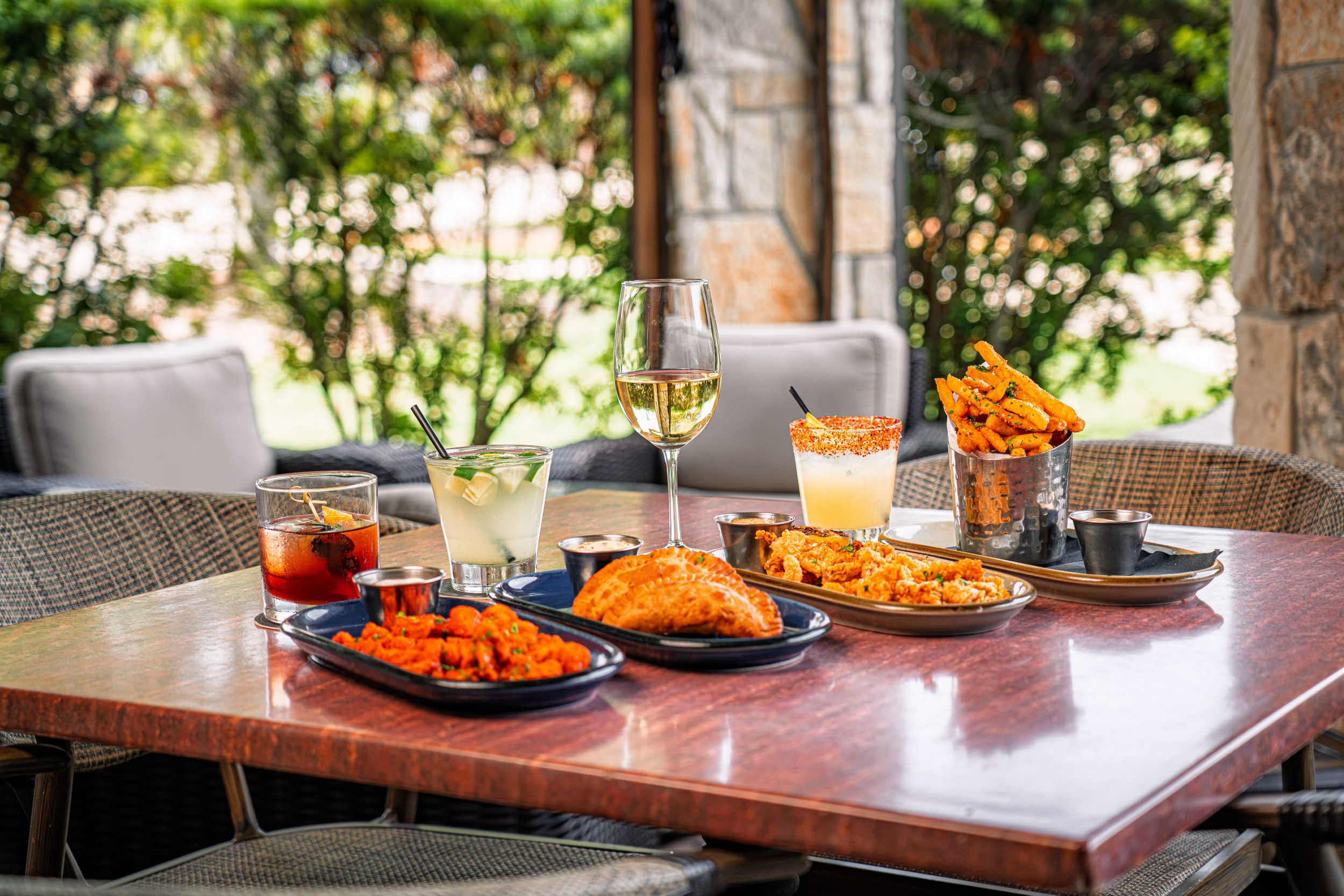 Buffalo chicken bites, Brazilian empanadas, calamari, and truffle fries spread across a patio table alongside four cocktails and a glass of white wine